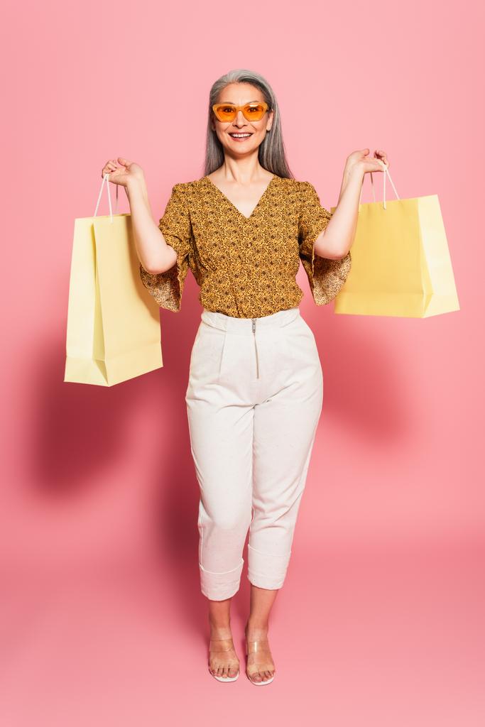 ピンクの背景にショッピングバッグを持つ流行の服で成熟したアジアの女性の完全な長さビュー - 写真・画像