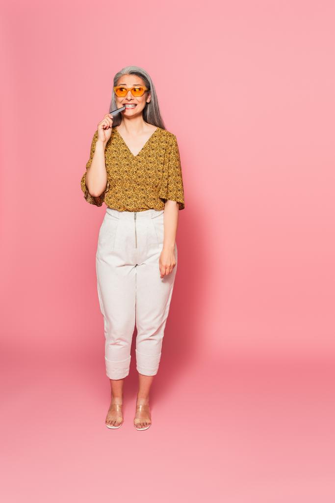 nervous asian woman in trendy clothes biting credit card while looking away on pink background - Photo, Image