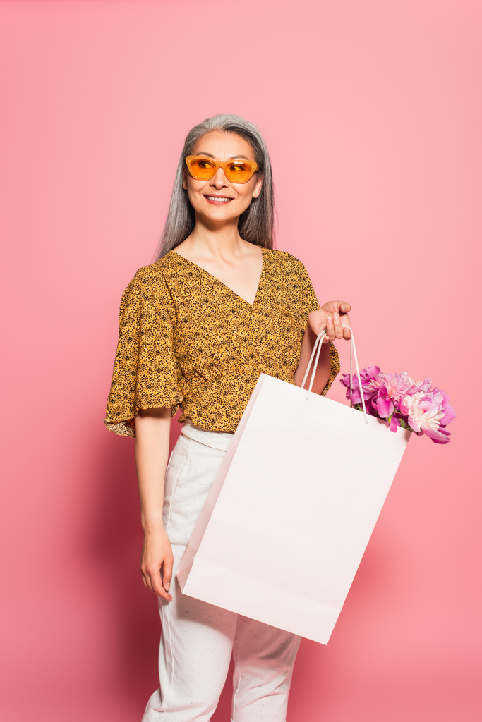elegante mujer de mediana edad con peonías en bolsa de compras blanca sobre fondo rosa - Foto, Imagen