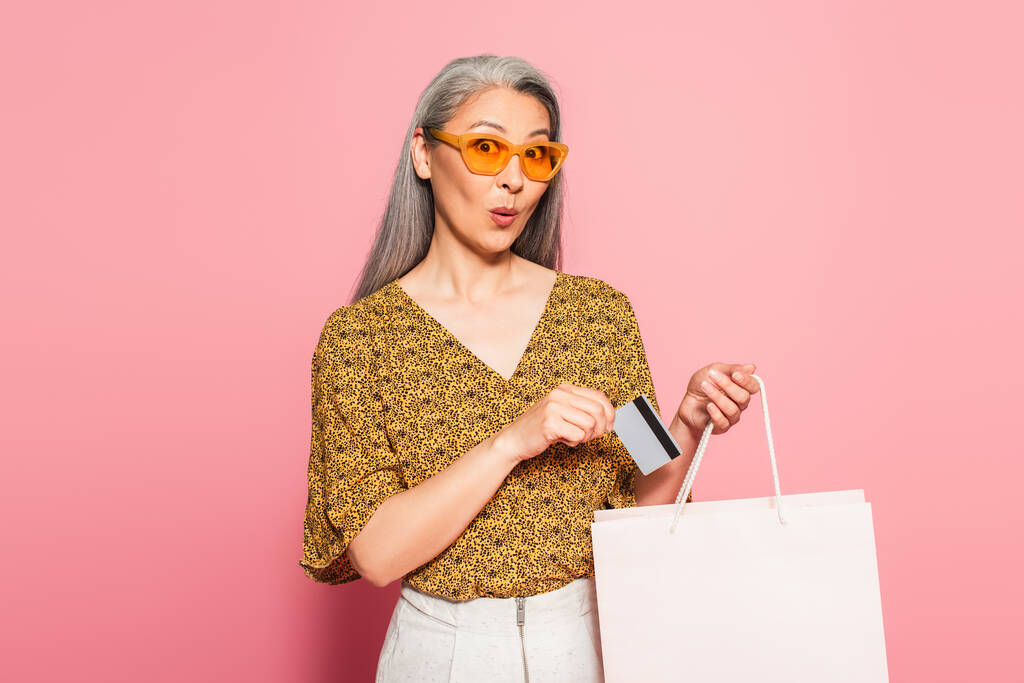 asombrado asiático mujer poniendo tarjeta de crédito en la bolsa de compras en rosa fondo - Foto, imagen