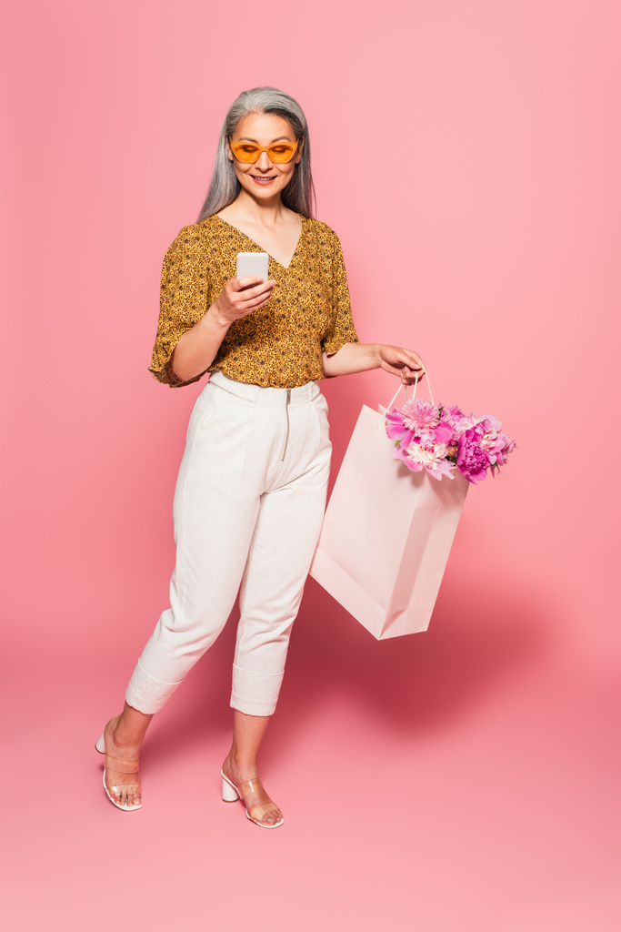 sorridente donna asiatica con peonie in shopping bag utilizzando smartphone su rosa  - Foto, immagini