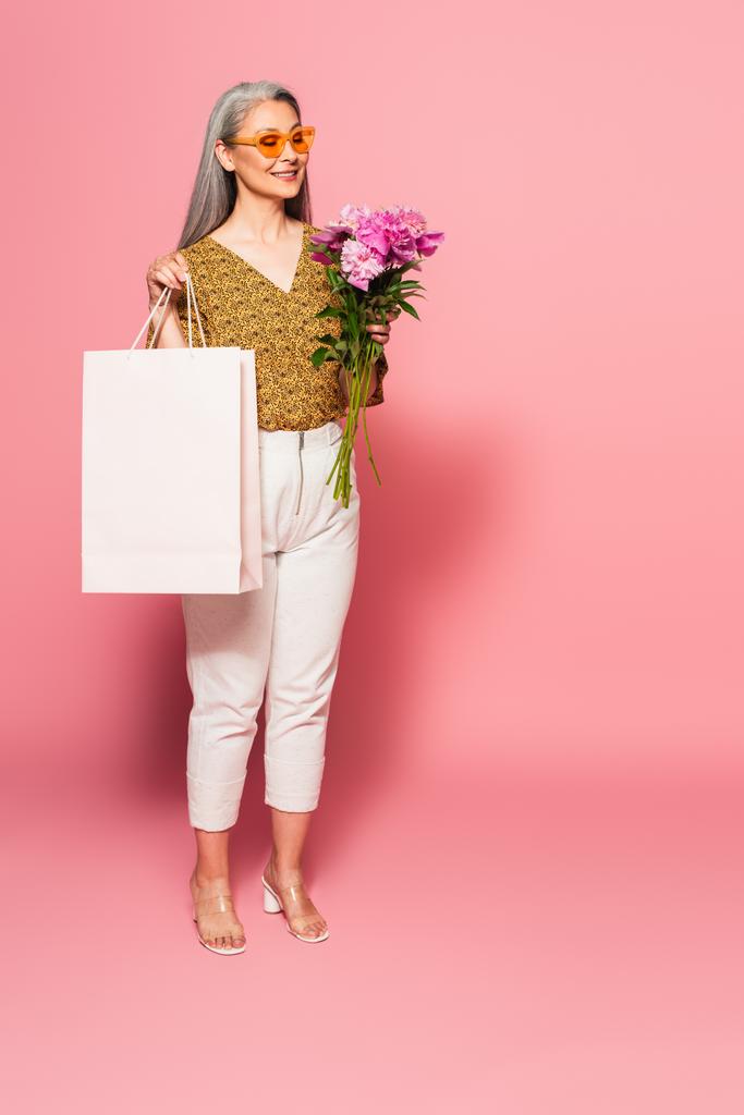 ピンクの上に牡丹とショッピングバッグを持つ幸せなアジアの女性の完全な長距離ビュー  - 写真・画像