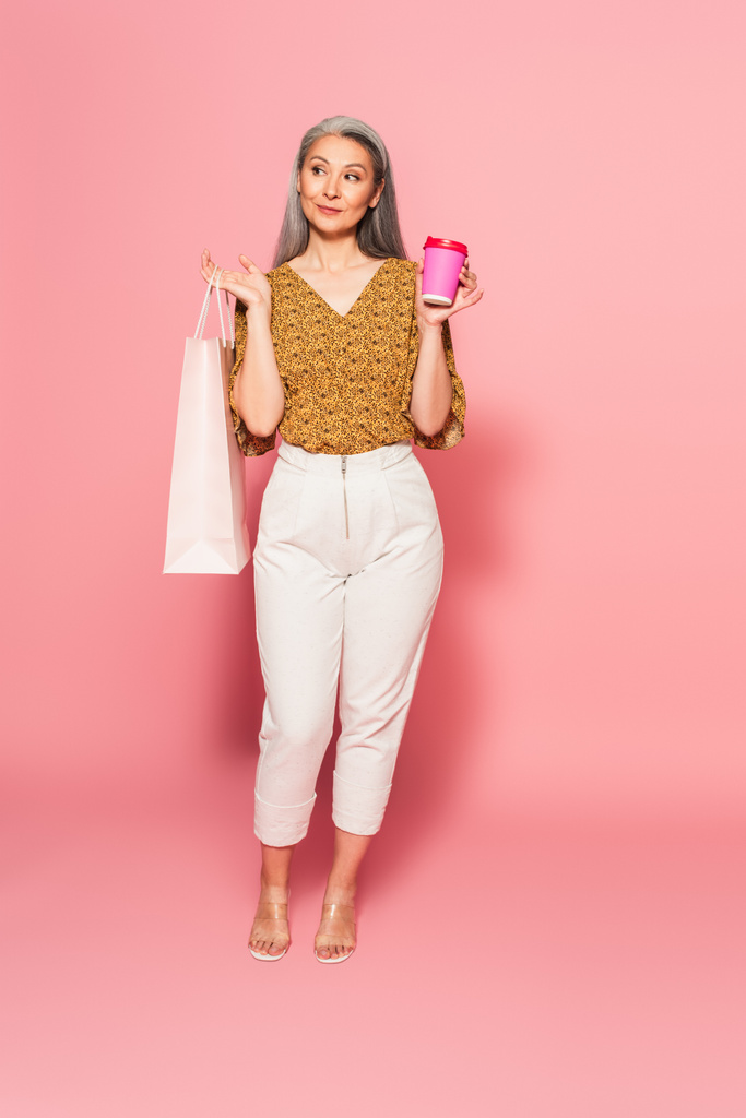 vista completa de la mujer asiática con taza de papel y bolsa de compras mirando hacia otro lado en rosa - Foto, imagen