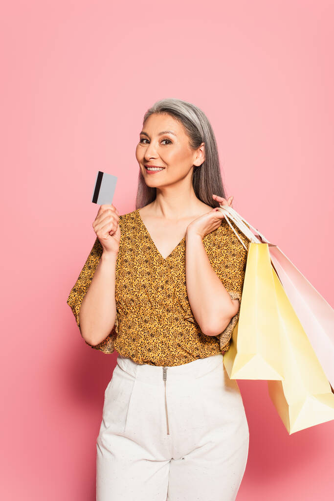 stylish asian woman with shopping bags showing credit card on pink background - Photo, Image