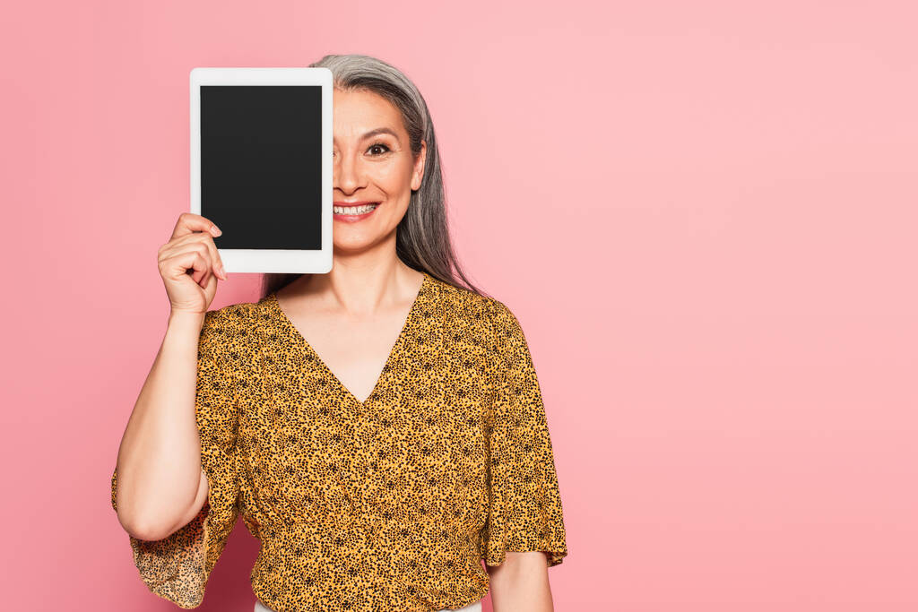 lächelnde asiatische Frau verdeckt Gesicht mit digitalem Tablet isoliert auf rosa - Foto, Bild