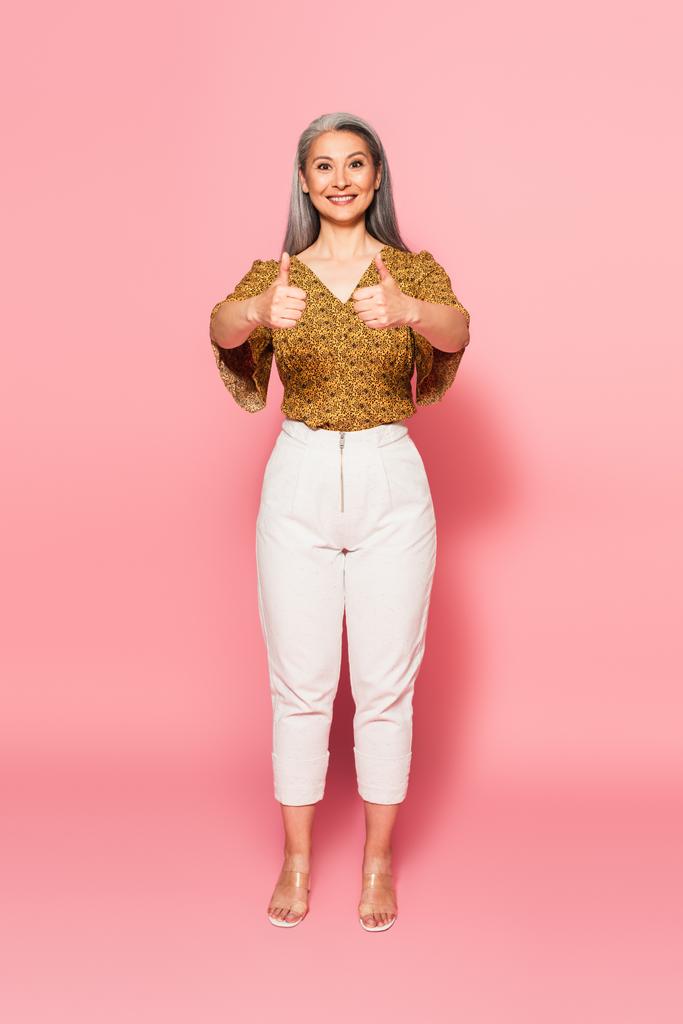 full length view of asian woman in pattern blouse and white pants showing thumbs up on pink  - Фото, изображение