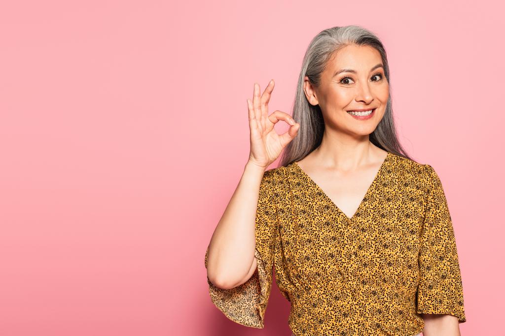 glücklich asiatische Frau in gelber Bluse mit Muster zeigt okay Geste auf rosa Hintergrund - Foto, Bild
