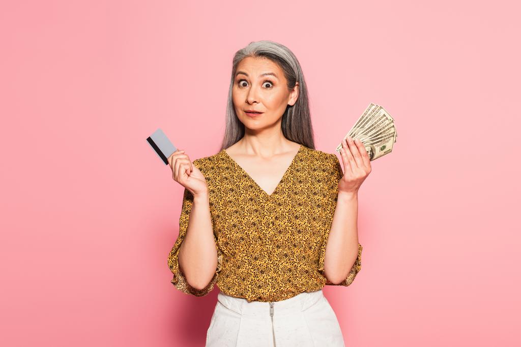 забавная азиатка с кредиткой и долларовыми банкнотами, смотрящая в камеру на розовом фоне - Фото, изображение