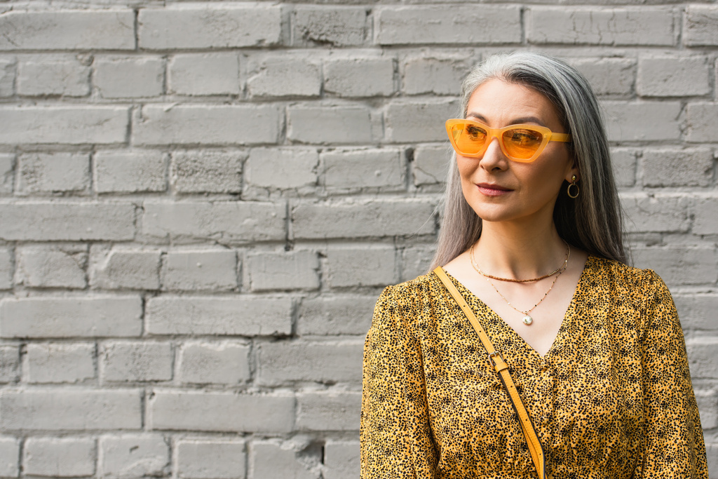 зрелая азиатка в стильных солнцезащитных очках, смотрящая в сторону кирпичной стены города - Фото, изображение
