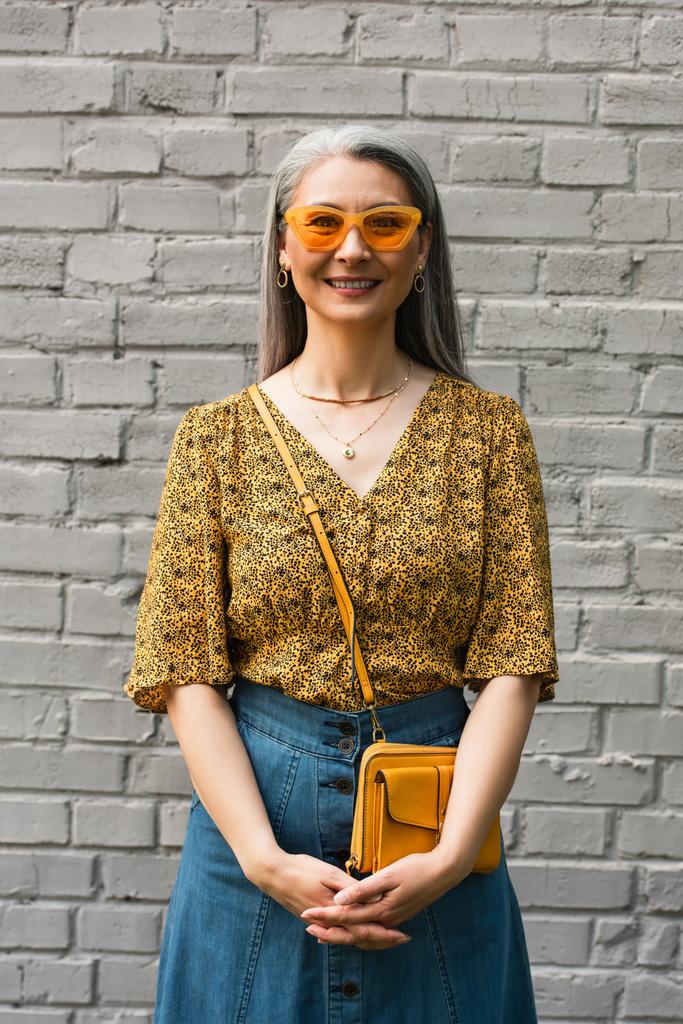 灰色のレンガの壁の近くにカメラを見て流行のブラウスと黄色のサングラスで幸せなアジアの女性 - 写真・画像