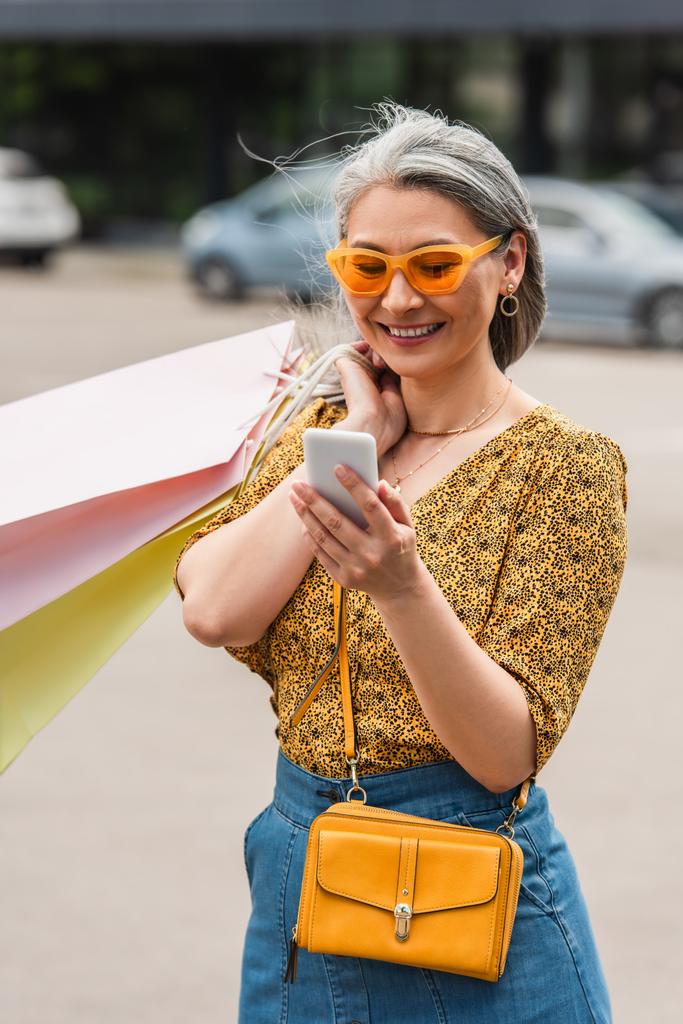 modna azjatycka kobieta z torbami na zakupy patrząc na telefon komórkowy na ulicy - Zdjęcie, obraz
