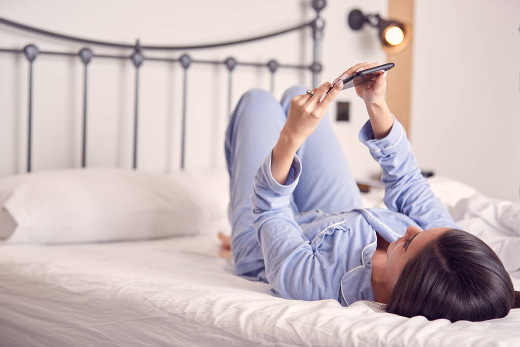 Mujer acostada en la cama con el teléfono móvil usando pijamas - Foto, imagen