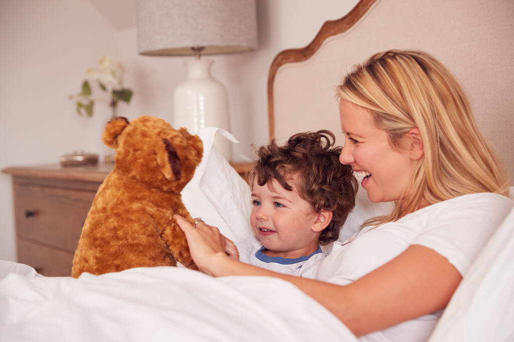 Mère et jeune fils portant des pyjamas dans la chambre à coucher ensemble jouant avec peluche peluche ours jouet - Photo, image