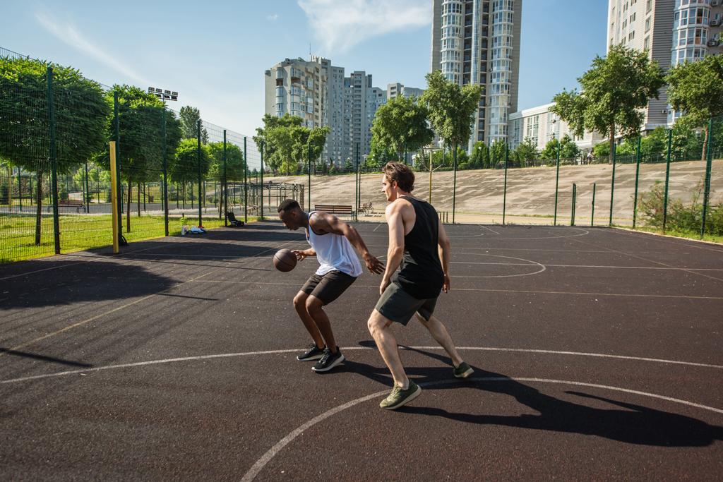 Vista laterale dell'uomo afroamericano con pallone da basket in esecuzione vicino amico sul parco giochi  - Foto, immagini