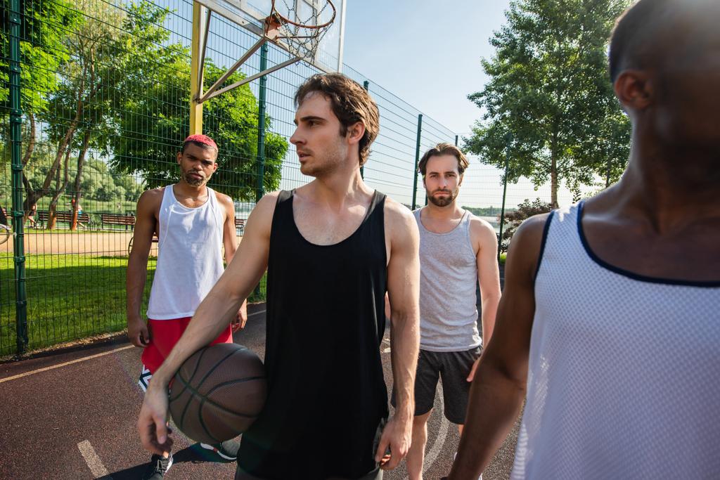 Νεαρός που κρατά μπάλα μπάσκετ κοντά σε πολυεθνικούς φίλους στην ύπαιθρο  - Φωτογραφία, εικόνα