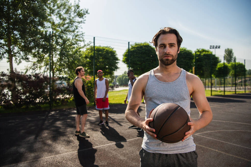 Hombre con pelota de baloncesto mirando a la cámara cerca de amigos interracial en la cancha  - Foto, Imagen