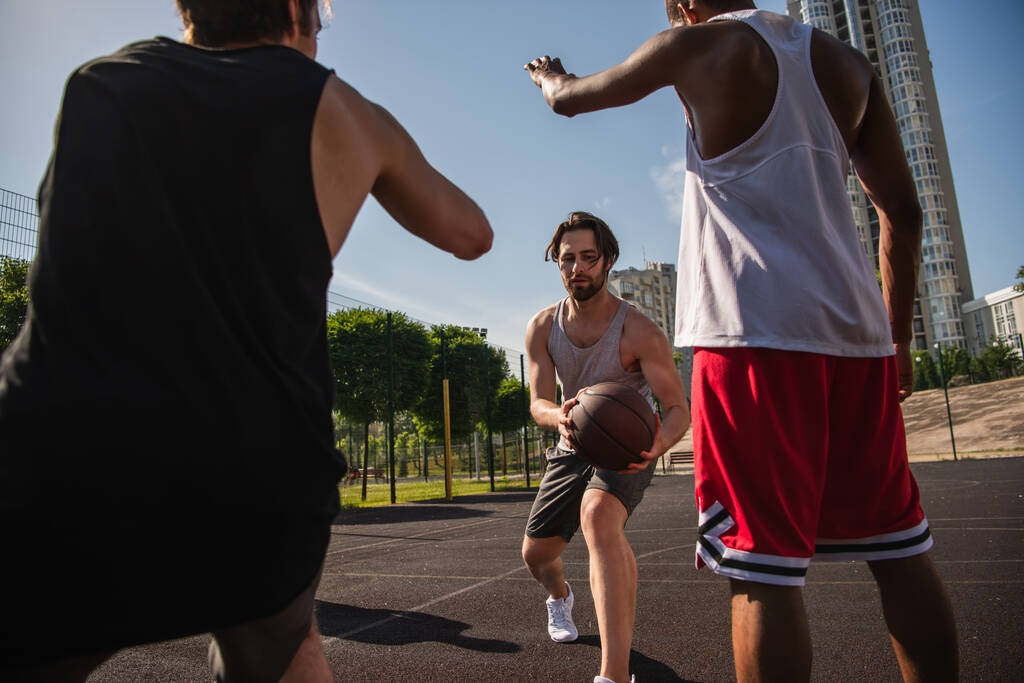 Νεαρός που κρατά μπάλα μπάσκετ ενώ παίζει με πολυεθνικούς φίλους στην ύπαιθρο  - Φωτογραφία, εικόνα