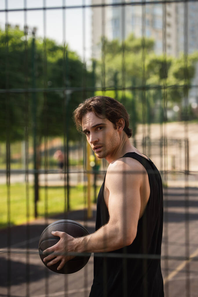 Mladý muž s basketbalovým míčem při pohledu na kameru v blízkosti plotu venku  - Fotografie, Obrázek