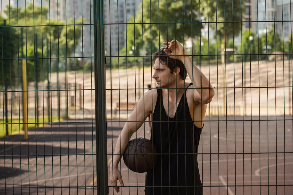 Boční pohled na sportovce s basketbalovým míčem stojící u plotu hřiště  - Fotografie, Obrázek