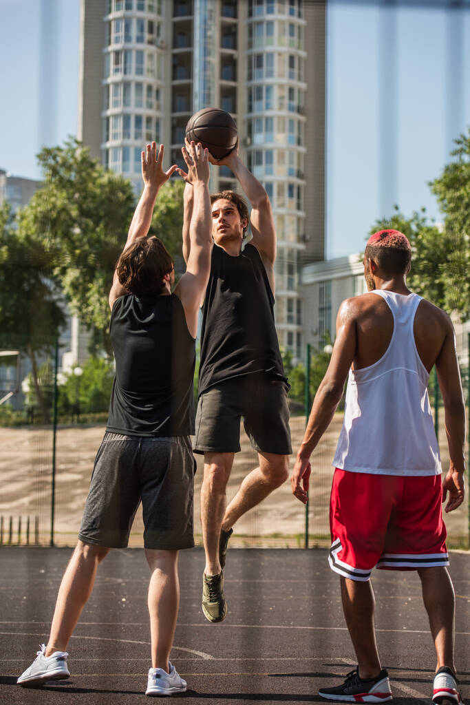 Sportowiec z piłką do koszykówki skacze w pobliżu międzyrasowych sportowców na świeżym powietrzu  - Zdjęcie, obraz