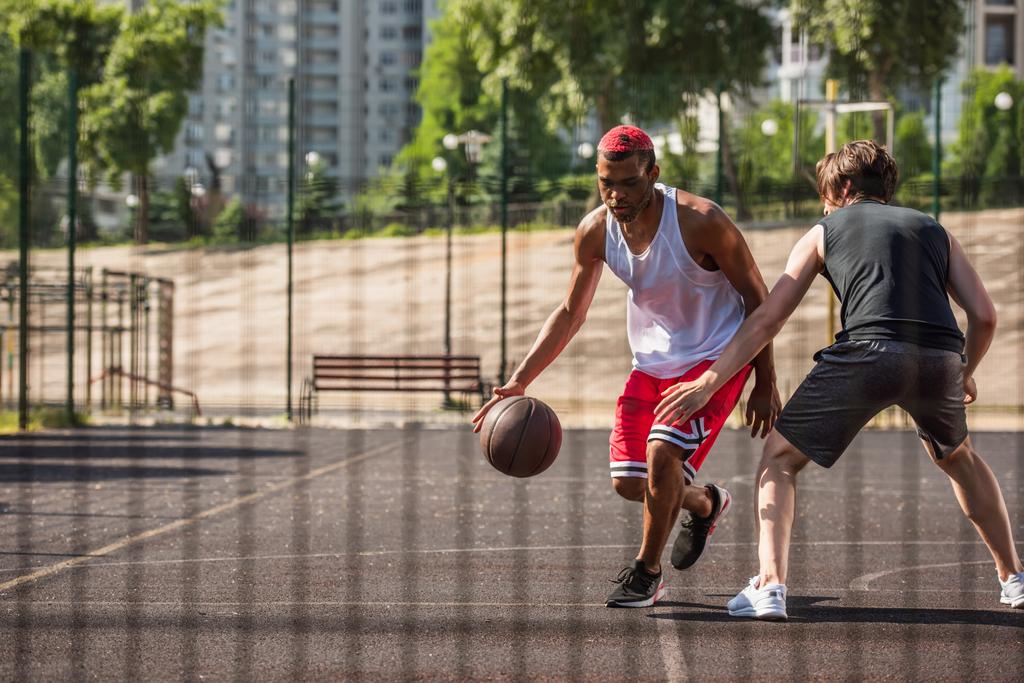 Uomo afro-americano che gioca a streetball vicino all'amico e alla recinzione offuscata  - Foto, immagini