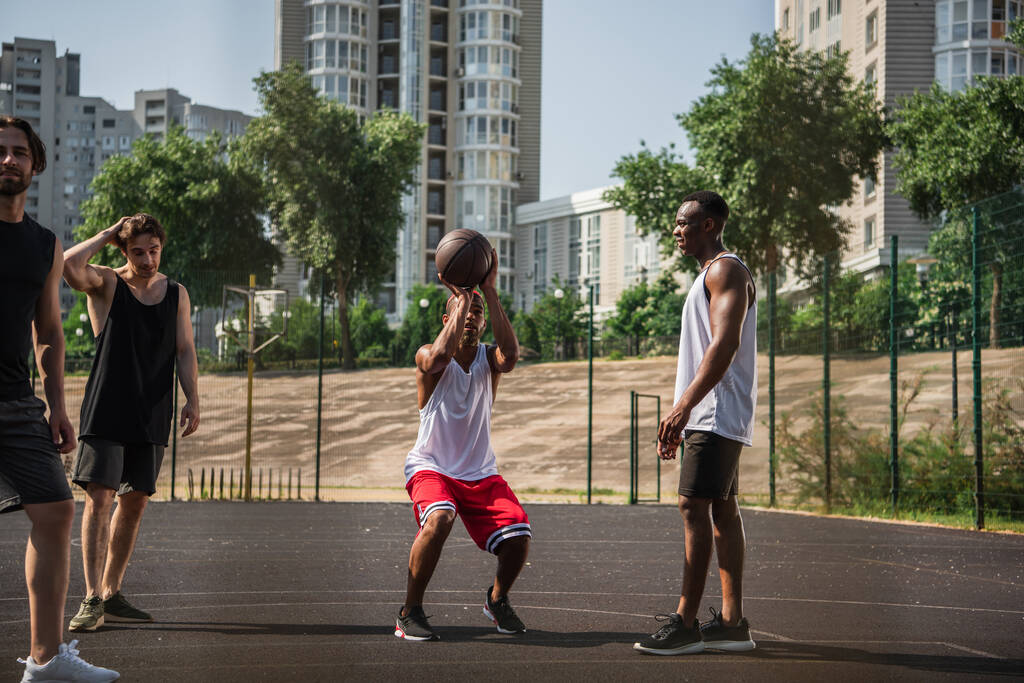 African american sportsman training with basketball ball near friends on playground on urban street  - Photo, Image