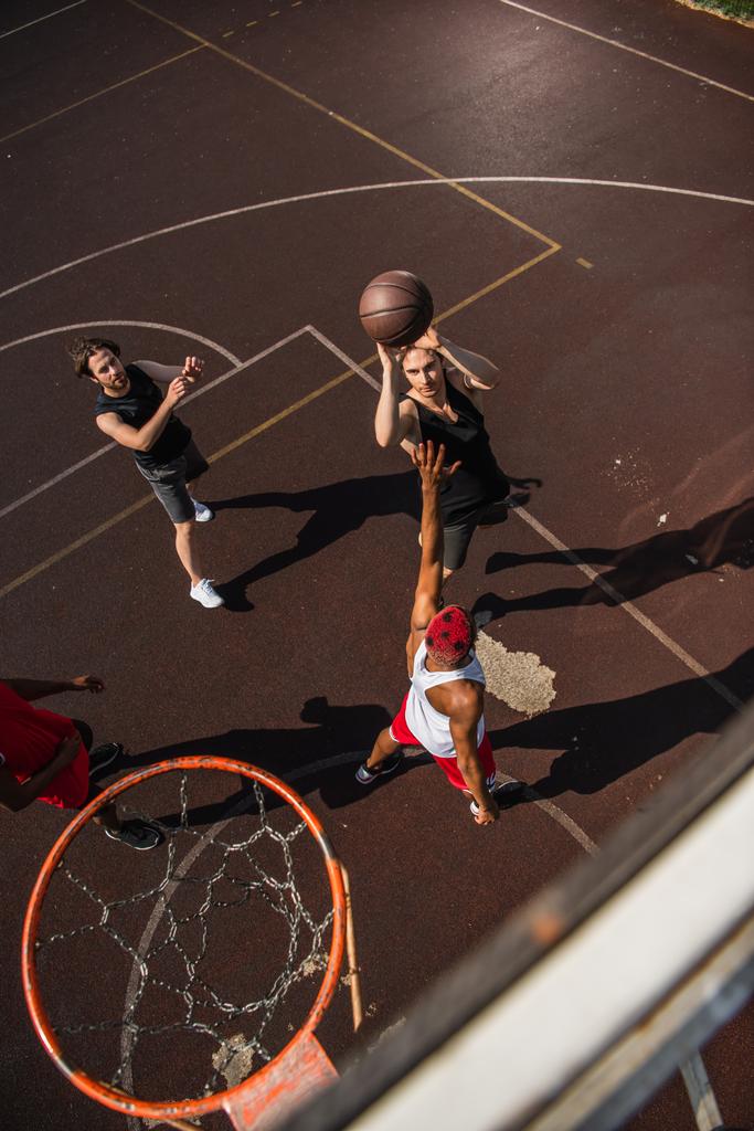 Bovenaanzicht van de mens met basketbal bal springen in de buurt van interraciale vrienden en hoepel buiten  - Foto, afbeelding