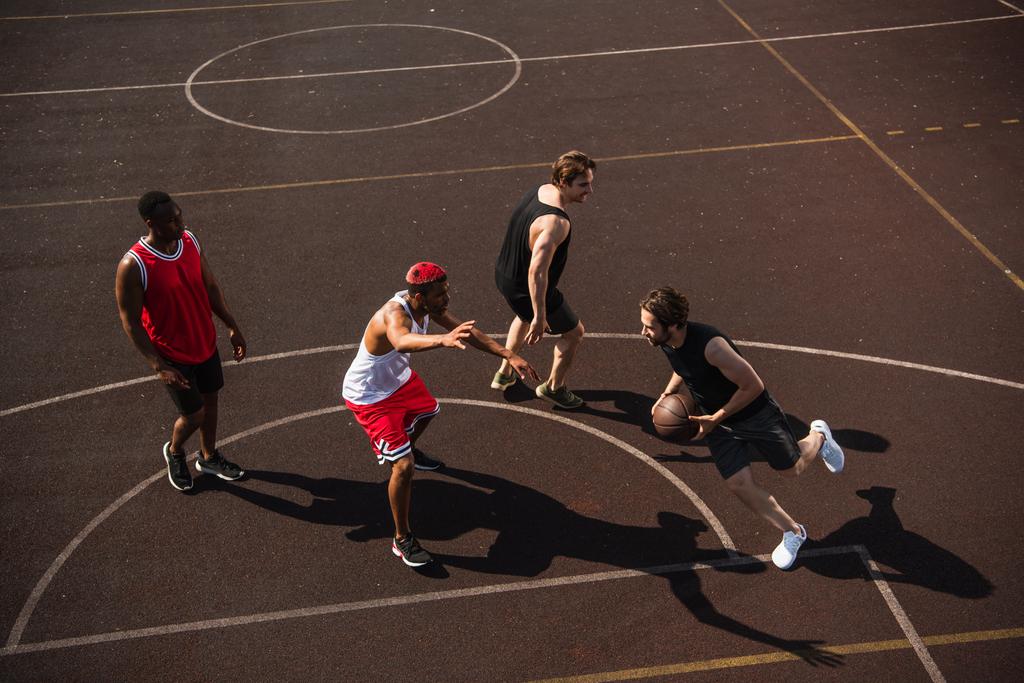 Άποψη υψηλής γωνίας του ανθρώπου με μπάλα μπάσκετ τρέχει στην παιδική χαρά κοντά σε πολυεθνικούς φίλους  - Φωτογραφία, εικόνα