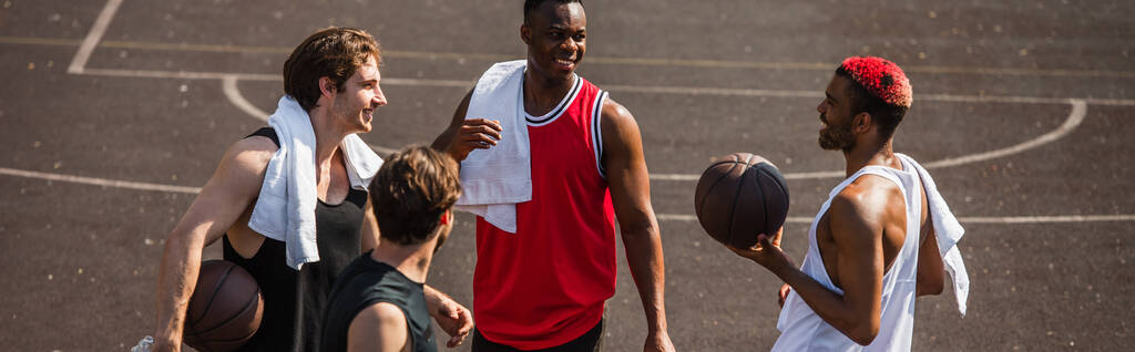 Positivo hombre afroamericano sosteniendo pelota de baloncesto cerca de amigos sonrientes con toallas al aire libre, pancarta  - Foto, imagen