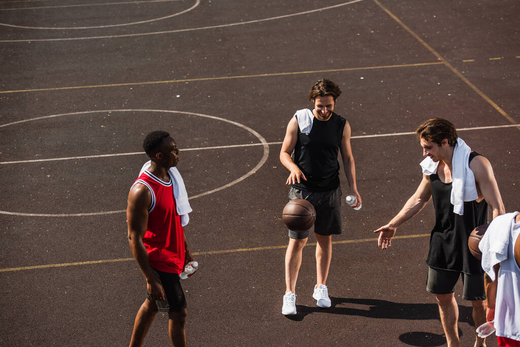 Hombre sonriente jugando baloncesto cerca de amigos interracial con toallas y botellas de agua en el patio de recreo  - Foto, Imagen