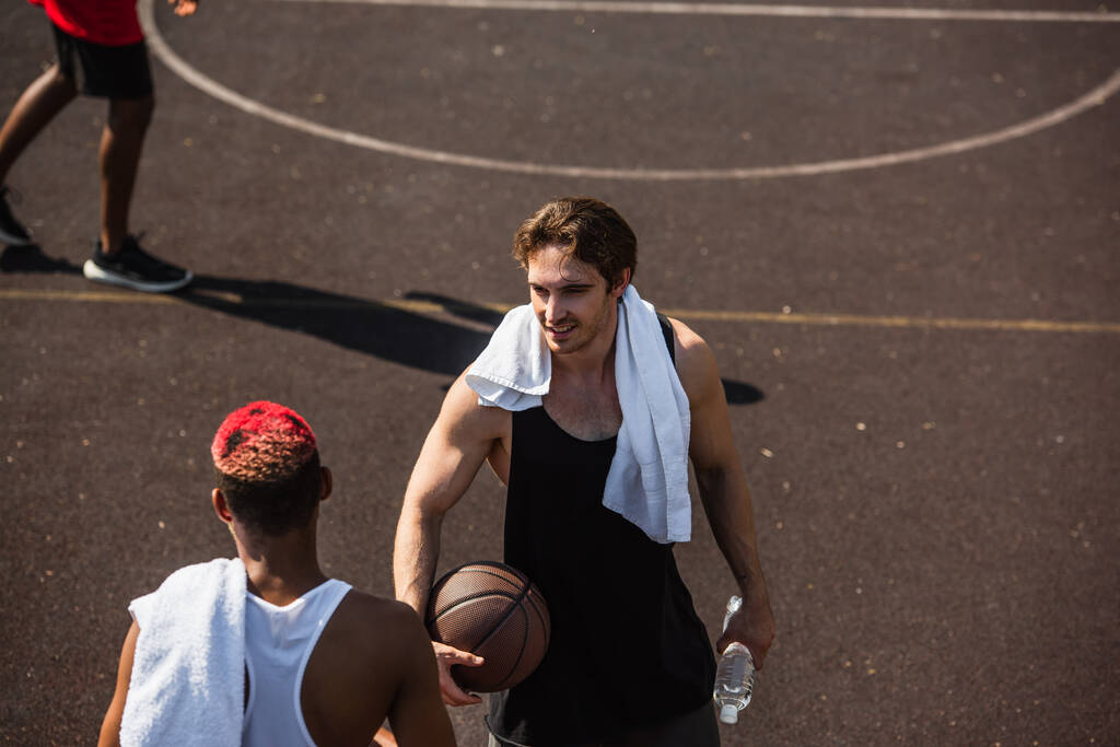 Vista de ángulo alto del hombre sonriente con toalla y pelota de baloncesto mirando a un amigo afroamericano  - Foto, imagen