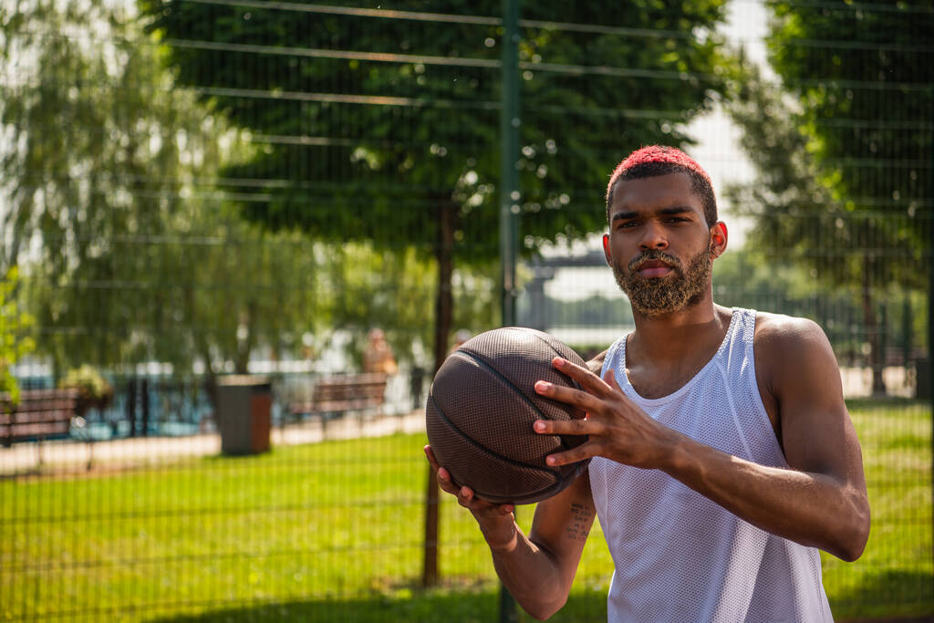 Αφροαμερικάνος αθλητής με μπάλα μπάσκετ κοιτάζοντας κάμερα σε εξωτερικούς χώρους  - Φωτογραφία, εικόνα