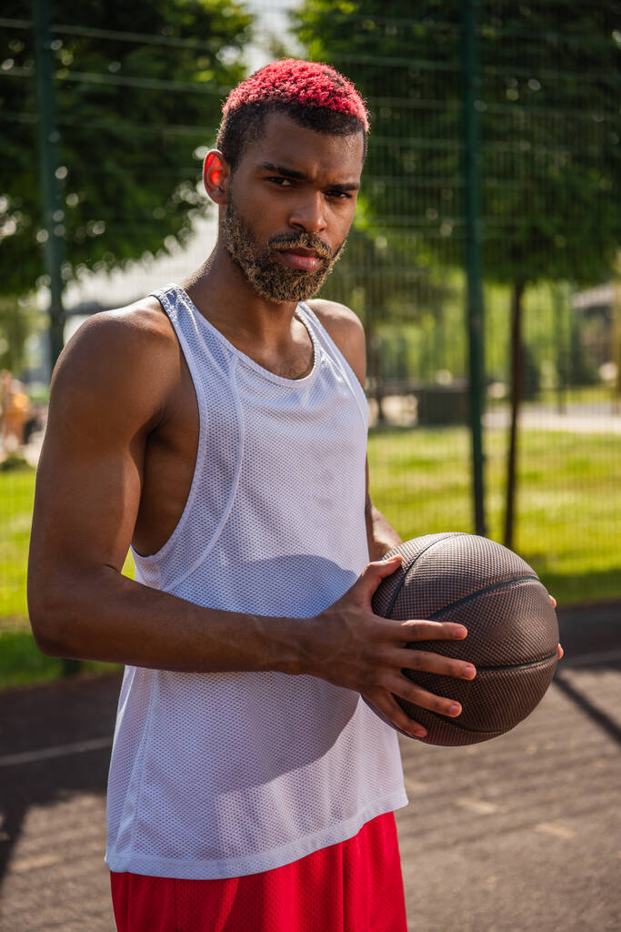 Siyah saçlı Afro-Amerikan sporcu basketbol topunu tutuyor ve dışarıda kameraya bakıyor.  - Fotoğraf, Görsel