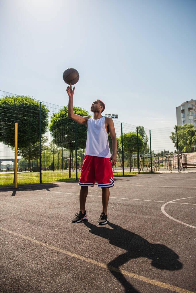 Widok z boku afrykański amerykański sportowiec trzymając piłkę do kosza na palcu na placu zabaw  - Zdjęcie, obraz