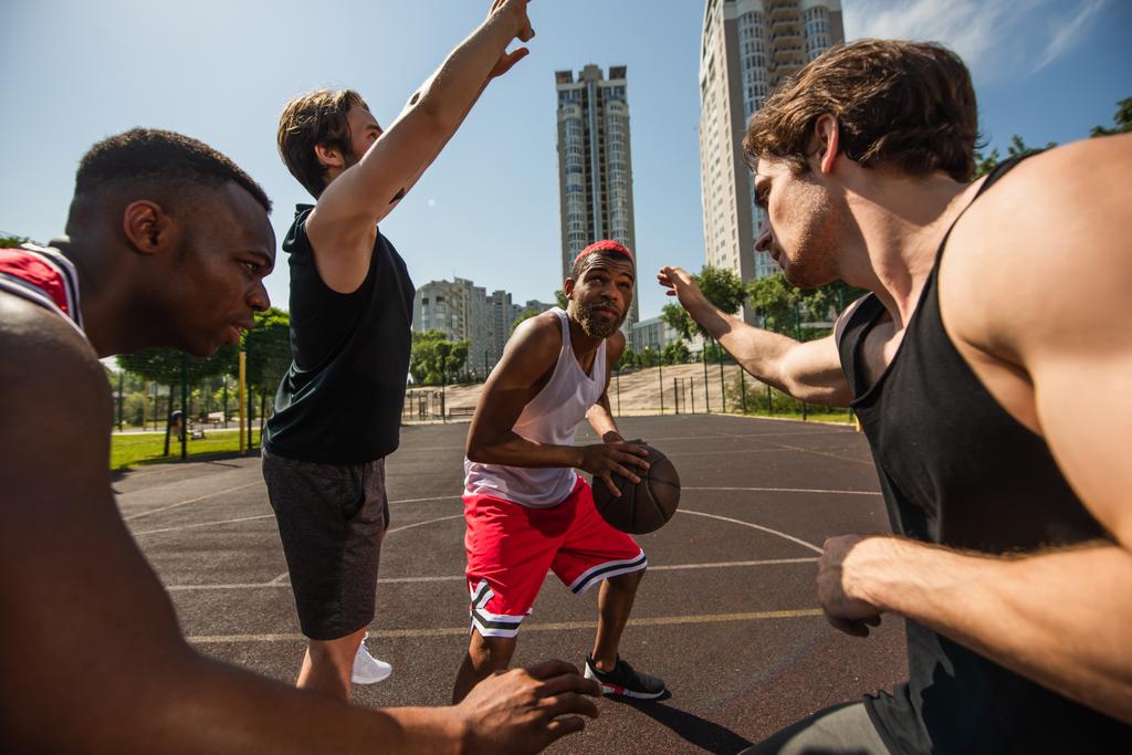 Deportista afroamericano sosteniendo pelota de baloncesto cerca de jugadores en la cancha  - Foto, Imagen