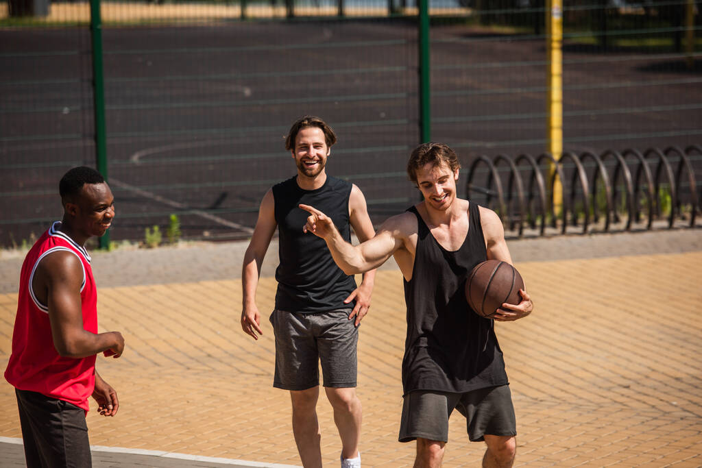 Lächelnder Sportler mit Basketballball zeigt mit dem Finger auf afrikanisch-amerikanischen Freund draußen  - Foto, Bild
