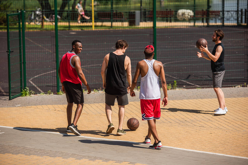 Fröhliche multiethnische Freunde mit Basketballbällen, die in der Nähe des Spielplatzes im Freien laufen  - Foto, Bild