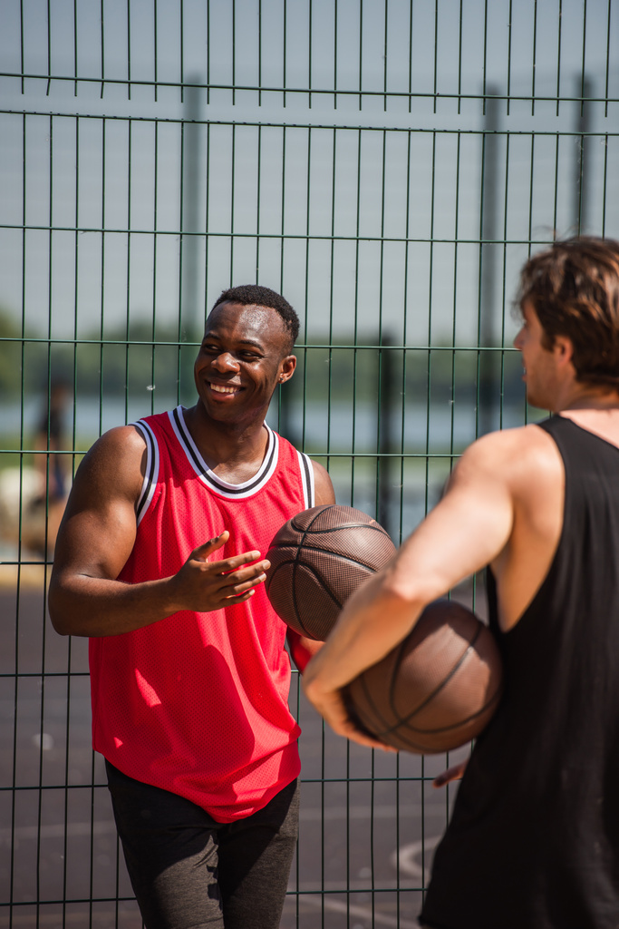 フェンスの近くにバスケットボールボールを保持する幸せなアフリカ系アメリカ人スポーツマンとぼやけた友人  - 写真・画像