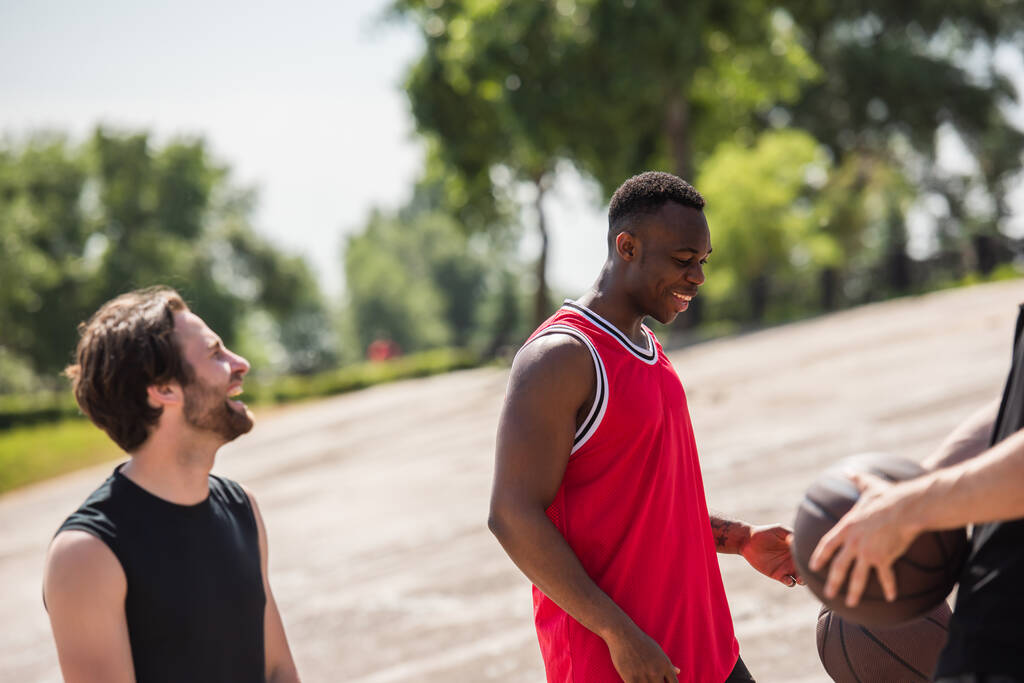 Улыбающийся африканский американец, стоящий рядом с друзьями с баскетбольными мячами на размытом переднем плане  - Фото, изображение