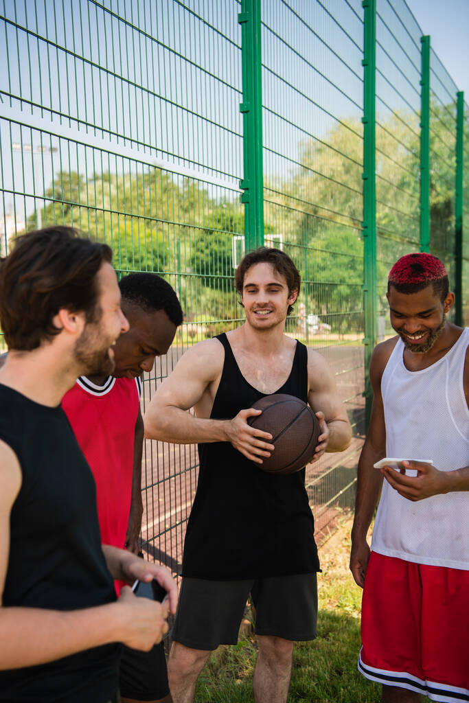 Χαμογελώντας άνθρωπος κρατώντας μπάλα μπάσκετ κοντά διαφυλετικούς φίλους με smartphones σε εξωτερικούς χώρους  - Φωτογραφία, εικόνα