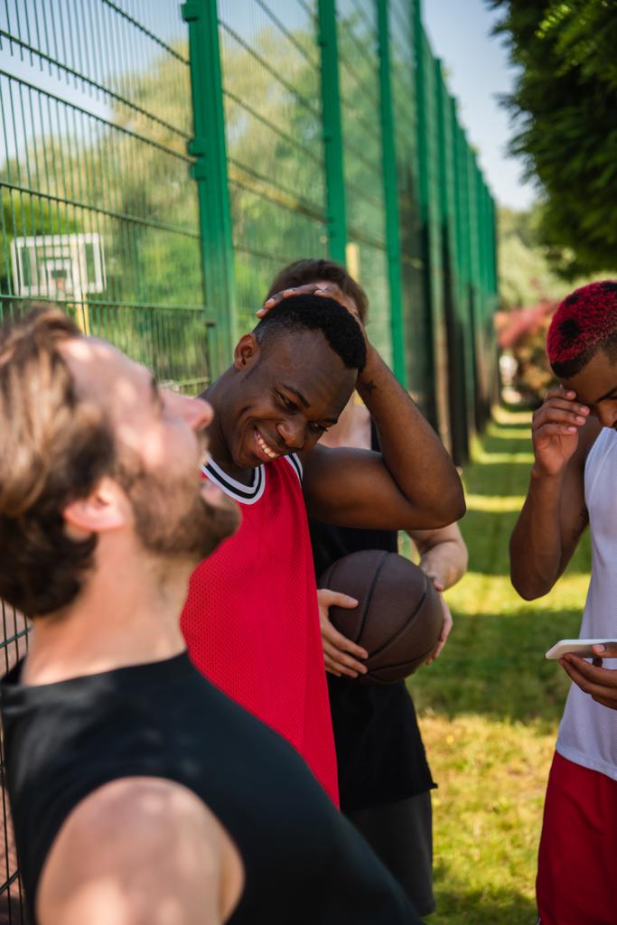Διαφυλετικοί παίκτες μπάσκετ με μπάλα και smartphone γέλιο σε εξωτερικούς χώρους  - Φωτογραφία, εικόνα
