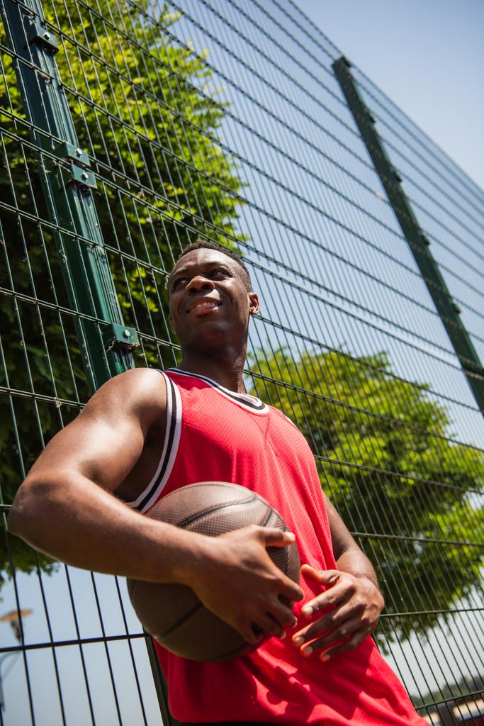 Χαμηλή γωνία άποψη του χαμογελαστού Αφροαμερικανός αθλητής κρατώντας μπάλα μπάσκετ κοντά φράχτη  - Φωτογραφία, εικόνα
