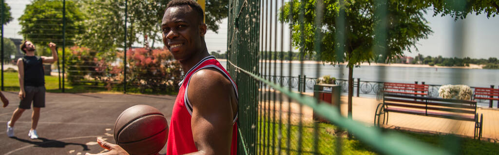 アフリカ系アメリカ人のスポーツマンを笑顔でバスケットボールボールを見てカメラ近くのぼやけたフェンス,バナー  - 写真・画像