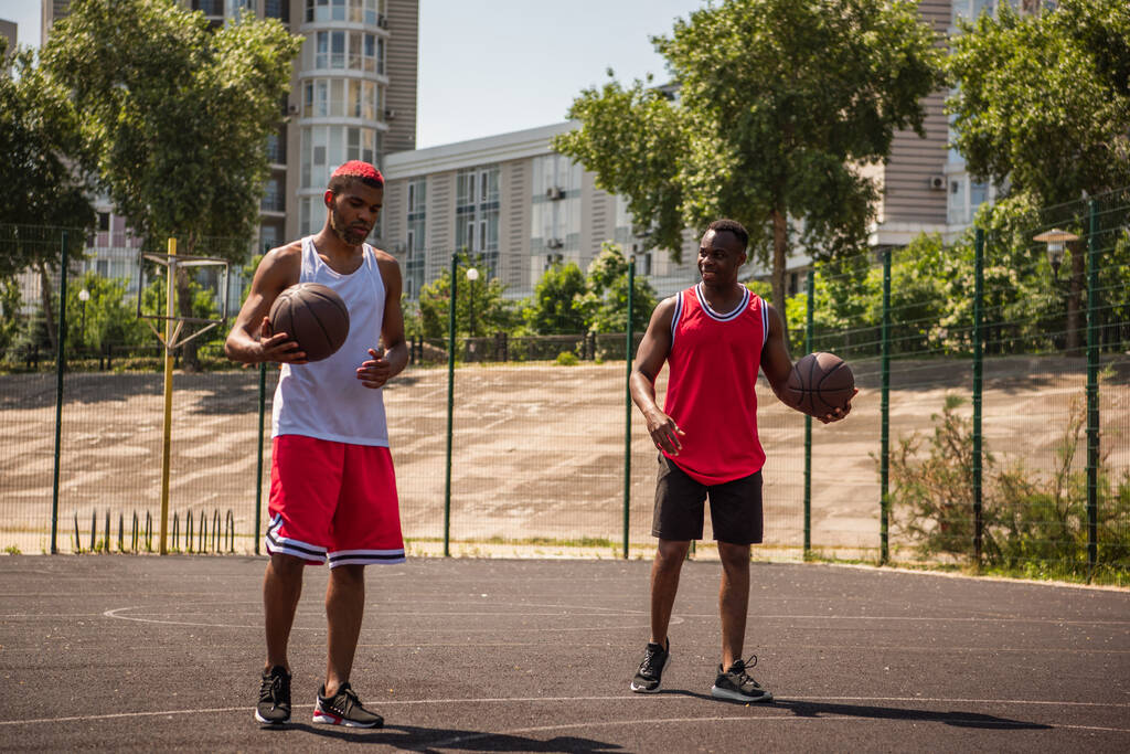 Χαμογελώντας Αφροαμερικανός αθλητής κοιτάζοντας φίλο με μπάλα μπάσκετ στο γήπεδο  - Φωτογραφία, εικόνα