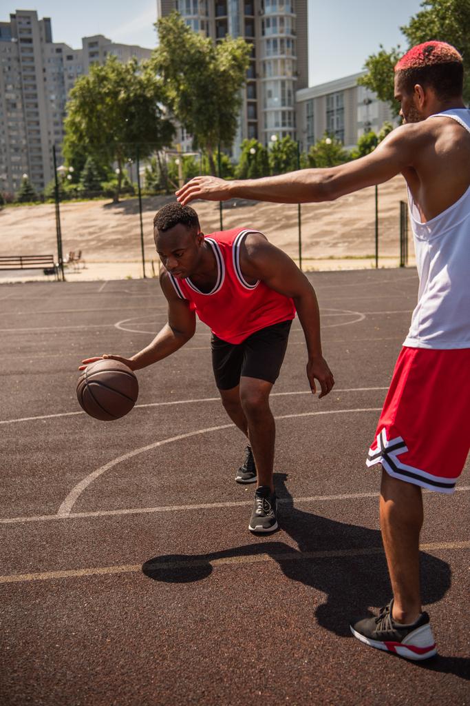 アフリカ系アメリカ人のスポーツマンがコートで友人の近くでバスケットボールをする  - 写真・画像