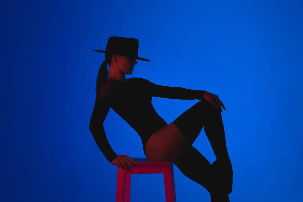 Επαγγελματίας χορευτής μπαλέτου στο καπέλο κάθεται σε ψηλή καρέκλα σε πολύχρωμο φόντο κάτω από βιολετί νέον φως. Αισθησιακή σέξι μπαλαρίνα που χορεύει με τα πόδια της σε κάλτσες. Γυναικεία μοιρολατρική στολή. - Φωτογραφία, εικόνα