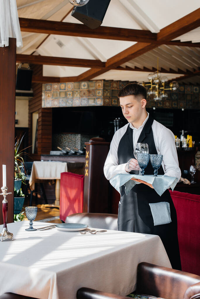 Ένας νεαρός σερβιτόρος με στυλάτη στολή ασχολείται με την εξυπηρέτηση του τραπεζιού σε ένα όμορφο γκουρμέ εστιατόριο. Δραστηριότητα εστιατορίου, του υψηλότερου επιπέδου. - Φωτογραφία, εικόνα