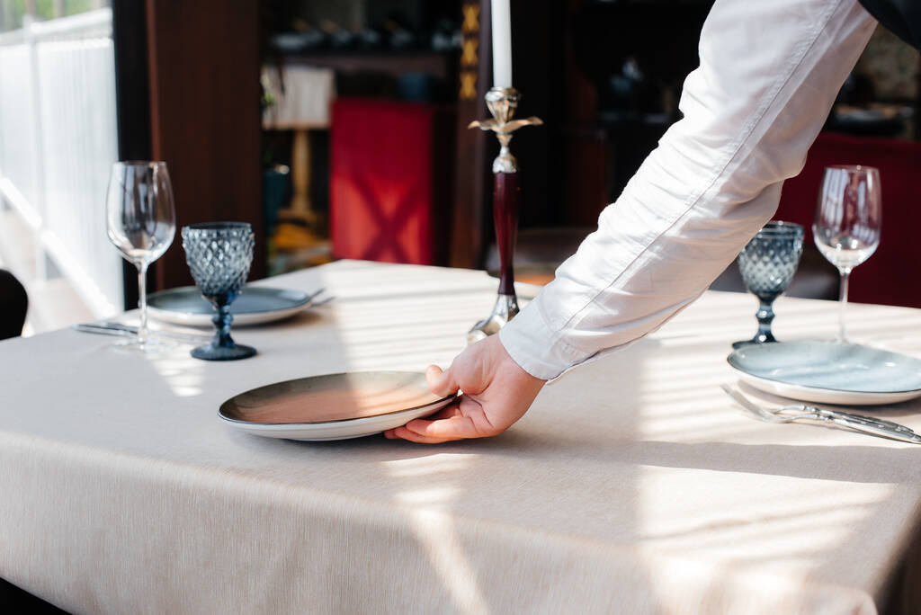 Een jonge ober in een stijlvol uniform serveert de tafel in een prachtig gastronomisch restaurant van dichtbij. Restaurant activiteit, van het hoogste niveau. - Foto, afbeelding