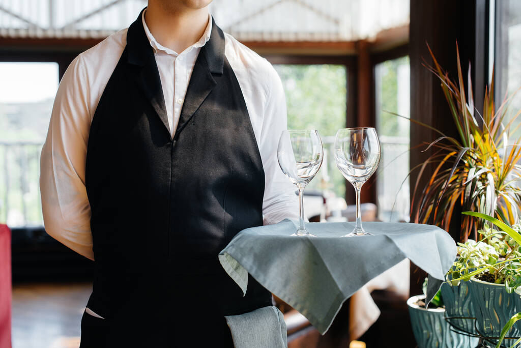 Un joven camarero con un elegante uniforme está de pie con vasos en una bandeja cerca de la mesa en un hermoso restaurante gourmet de cerca. Actividad del restaurante, del más alto nivel. - Foto, imagen