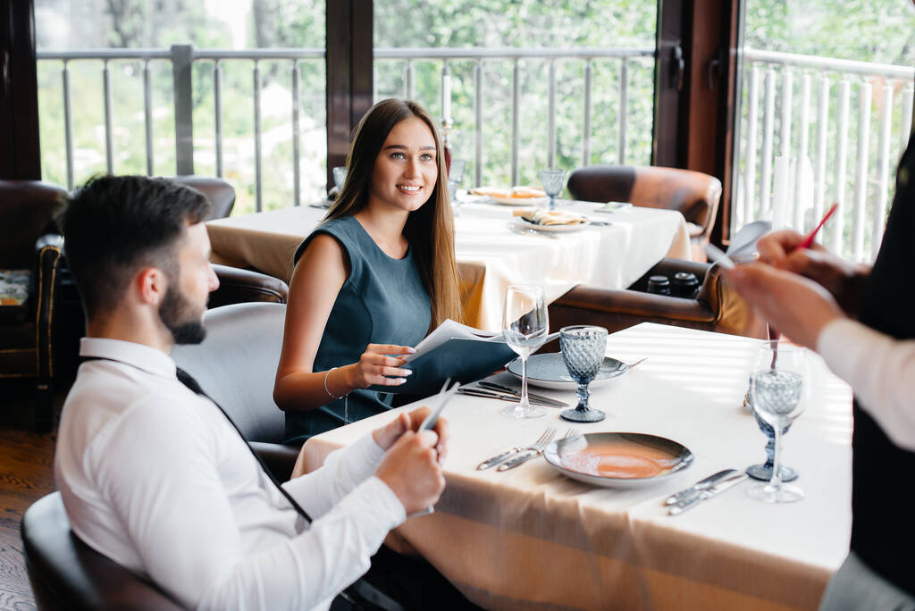 Un camarero elegante sirve a una joven pareja que vino en una cita en un restaurante gourmet. Servicio al cliente en el catering. - Foto, Imagen